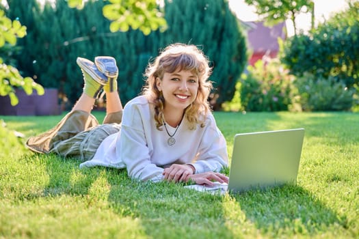 Teenage girl lying on grass using laptop, looking at camera. Female teenager student 16, 17 years old lying on lawn, technologies for studying leisure communication shopping in backyard in garden