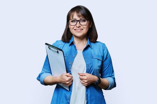 Middle aged business woman with clipboard looking at camera on white studio background. Mature smiling female teacher agent auditor psychologist supervisor posing with papers documents contracts