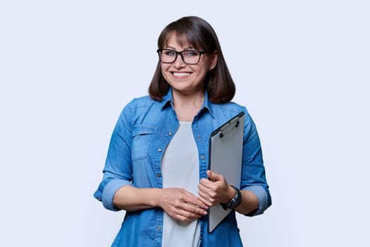 Middle aged business woman with clipboard looking at camera on white studio background. Mature smiling female teacher agent auditor psychologist supervisor posing with papers documents contracts