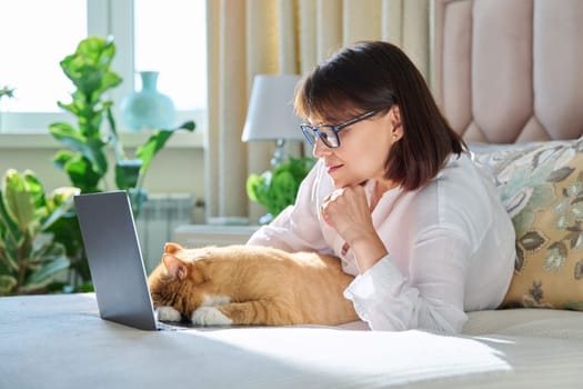 Middle aged woman at home on couch with laptop and cat. Mature 40s female looking at camera, on bed with pet. Work at home, technology, leisure, freelancing, lifestyle, people and animals concept