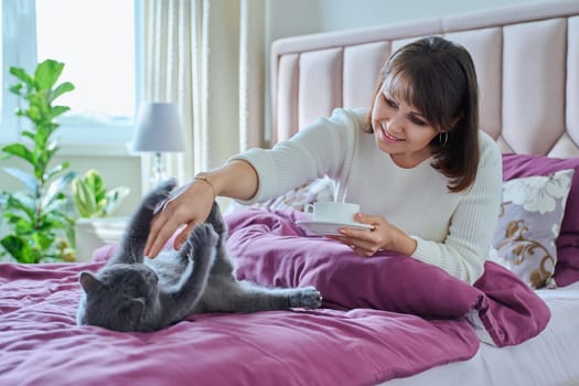 Middle-aged cheerful woman with cup of morning coffee in hand playing with cat in bed. Mature female owner touching pet, lifestyle friendship fun, people animals concept