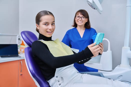 Happy young teenage female with mirror sitting in dentist chair smiling looking at healthy teeth, with dentist doctor in office. Dentistry, hygiene, treatment, dental health care concept