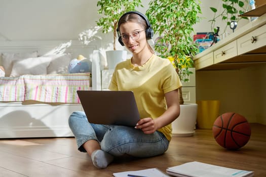 Teenage girl with glasses studying at home using laptop. Female with headphones and textbooks sitting on floor in room, looking at camera. Education, adolescence, high school concept