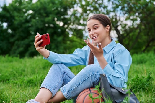 Teenage female student resting sitting on grass using smartphone for video call chat conference. Attractive teen girl on lawn with backpack. High school applications technology lifestyle adolescence