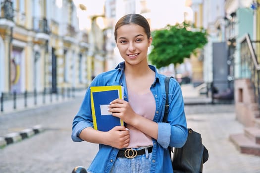 Outdoor portrait of smiling teenage female student looking at camera in city. Girl with backpack with books and notebooks in hands. Education, knowledge, high school, adolescence concept