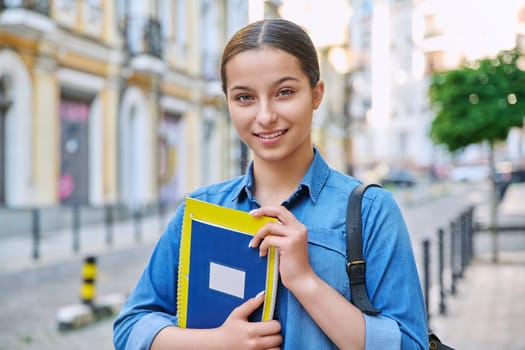 Outdoor portrait of smiling teenage female student looking at camera in city. Girl with backpack with books and notebooks in hands. Education, knowledge, high school, adolescence concept