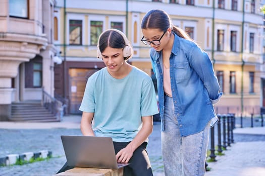 Teenage male and female using laptop for study, leisure, outdoor on city street. Guy and girl teenagers looking together at aptop screen. Lifestyle, technology, youth, education, city life concept