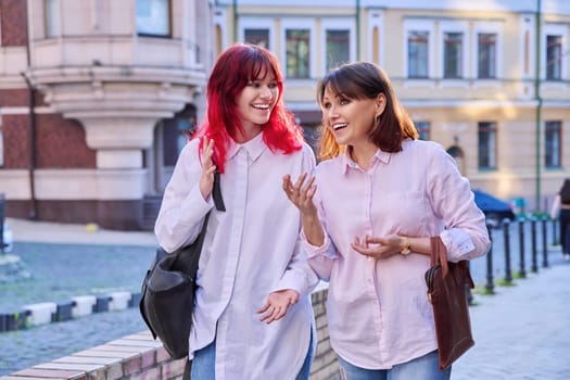 Two generations, smiling joyful happy mother and stylish attractive teenage daughter, walking talking together along city street. Family communication parent child teenager lifestyle leisure people