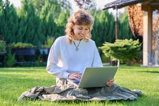 Teenage girl sitting on the grass using a laptop. Female teenager student 17, 18 years old sitting on the lawn, technologies for studying leisure communication shopping in backyard in garden