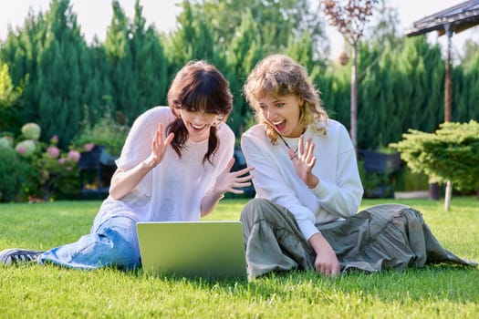 Young teenage female student friends sitting on grass with laptop. Teenagers rejoicing laughing and having fun together. Friendship, youth, technology, high school, college, lifestyle concept