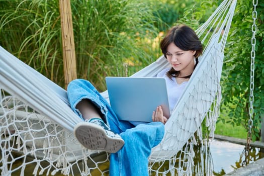 Teenage girl relaxing in hammock using laptop for leisure study. Adolescence, students, high school, technology, lifestyle, youth concept