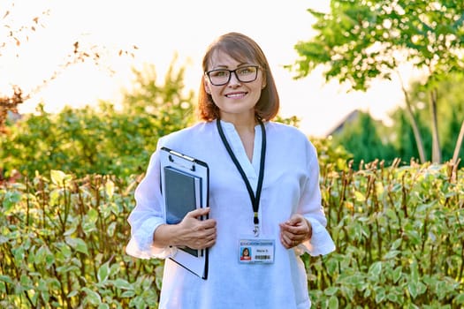 Outdoor portrait of female teacher with clipboard badge. Middle aged female mentor, educator, psychologist, social worker, counselor smiling looking at camera