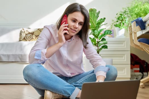 Young smiling woman sitting on floor at home talking on mobile phone using laptop. Education, study, remote online work, freelancing, youth concept