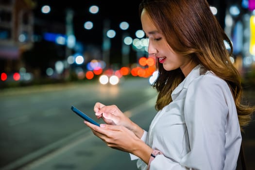 Portrait of a Young Asian woman on the streets of downtown. She using mobile app device on smartphone in downtown city street, with illuminated busy city traffic scene during rush hour.