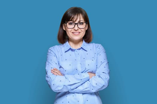 Middle aged smiling business woman over blue color background. Confident 40s female looking at camera with crossed arms. Teacher, psychologist, profession, psychology therapy, success, work, staff