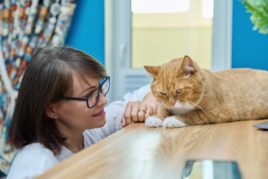 Middle aged woman talking with ginger pet cat, home interior background. Friendship, love, care, leisure, lifestyle, animals people house concept