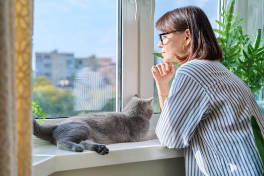 Mature smiling woman looking out window at home with relaxed cat lying on windowsill enjoying warmth of sun. Lifestyle home comfort joy happiness, animals pets, middle aged people concept