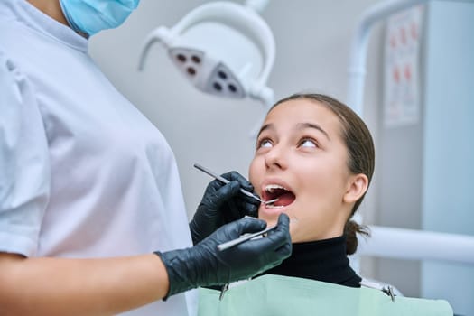 Young teenage female at dental checkup in clinic. Teenage girl sitting in chair, doctor dentist with tools examining patient's teeth. Adolescence, hygiene, dentistry, treatment, dental health care