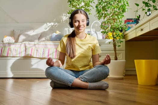 Teenage girl in headphones relaxing meditating sitting in lotus position at home on floor, happy adolescent teen female looking at camera. Recreation, lifestyle, adolescence, lifestyle, youth concept