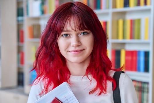 Portrait of teenage female student looking at camera in library. Smiling beautiful girl teenager with trendy red dyed hair holding notebook. College, university, education, knowledge, youth concept