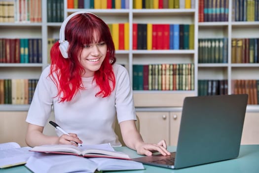 Teenage female student studying in library. Girl in headphones using laptop, looking at screen, watching webinar, making notes in notebook. Youth, technology, education, college, university concept