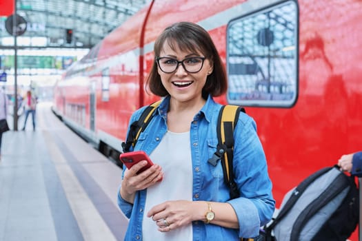 Woman waiting for railway public electric transport on platform of city station. Female with smartphone using banking app to buy online ticket payment, online timetable and route service, technology