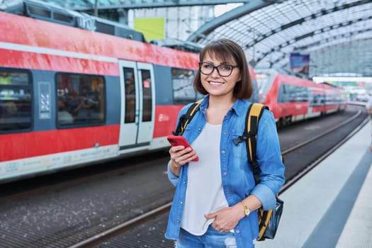 Woman waiting for railway public electric transport on platform of city station. Female with smartphone using banking app to buy online ticket payment, online timetable and route service, technology
