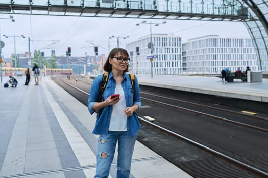 Woman waiting for railway public electric transport on platform of city station. Female with smartphone using banking app to buy online ticket payment, online timetable and route service, technology