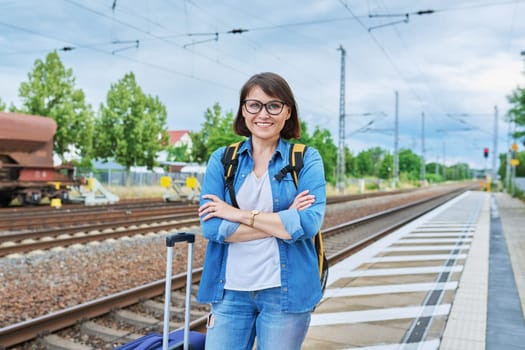 Middle aged woman with backpack with suitcase on outdoor platform of railway station looking at camera. Transport, luggage, journey, vacation, tourism, people concept