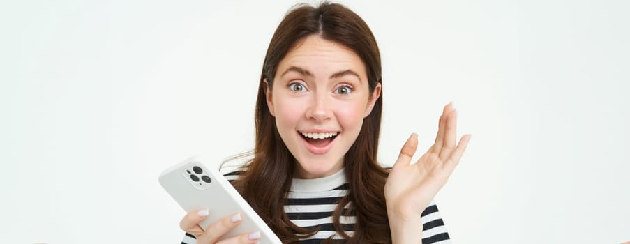 Portrait of happy, surprised young woman, holding mobile phone, showing amazed reaction to great news, standing with smartphone over white background.