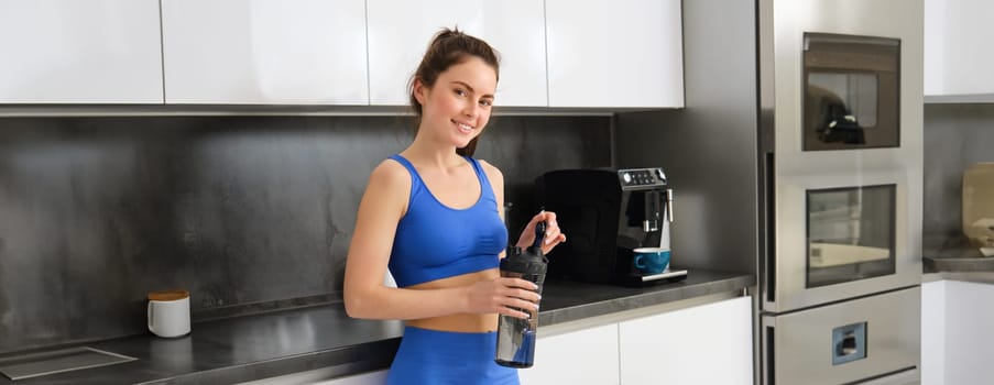 Portrait of beautiful fitness instructor, holding water bottle, drinking protein and smiling, standing near kitchen counter.