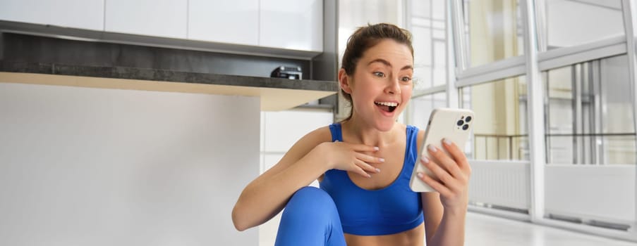 Indoor shot of surprised, happy young sportswoman looking at smartphone with smile and excitement, reading great news on phone message, sitting in blue sportsbra and leggings.