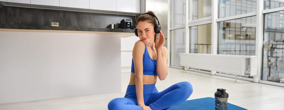 Young woman, fitness instructor doing workout at home, practice yoga in wireless headphones on rubber mat.