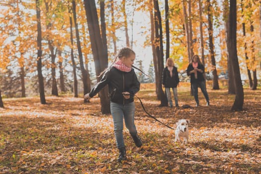 Little girl running with her dog jack russell terrier among autumn leaves. Mother and grandmother walks behind.