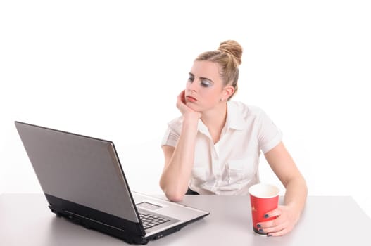 Young woman with blond hair wearing a business suit and has bored in the office. She sits far succeeded with a coffee cup in front of her computer keyboard, isolated against white background.