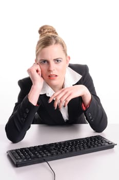 Young woman with blond hair wearing a business suit and has frustrated the computer. She sits at her keyboard and looks annoyed at the camera, isolated against white background.