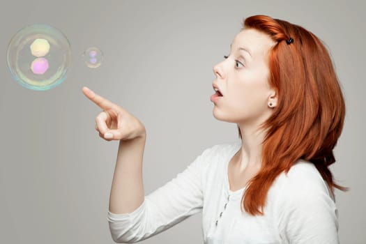 young beautiful red haired girl look to soap bubbles and wondering in front of grey background