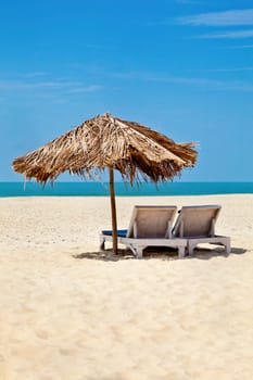 A generic vertical portrait of a tropical deserted beach with loungers under a coconut leaf parasol. Blue sky, ocean, gentle surf sound and clean sandy beach. Location, Goa India