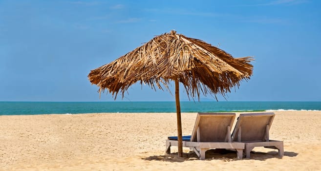 Panorama of a generic tropical deserted beach with loungers under a coconut leaf parasol. Blue sky, ocean, gentle surf and clean sandy beach. Location Uttorda Beach, Goa India