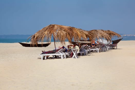 Landscape of secluded beach with day trippers and holidaymakers basking in the sun under the shade of coconut parasols. White sand, blue sky, sea, surf and fishing boats for a backdrop. Generic tropical shot location, Goa India