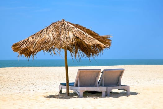 A generic tropical deserted beach with loungers under a coconut leaf parasol. Blue sky, ocean, gentle surf sound and clean sandy beach. Location, Uttorda Beach, Goa India
