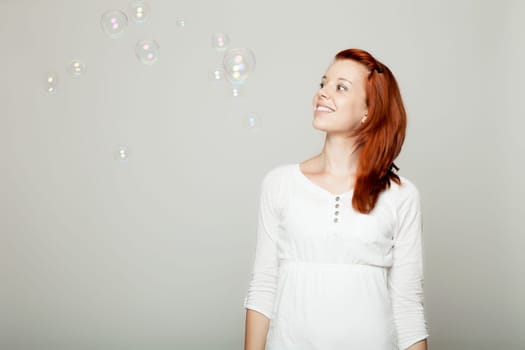 young beautiful red haired girl look to soap bubbles and dream in front of grey background