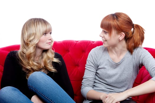 young beautiful blond and red haired girls talking and sitting on red sofa in front of white background