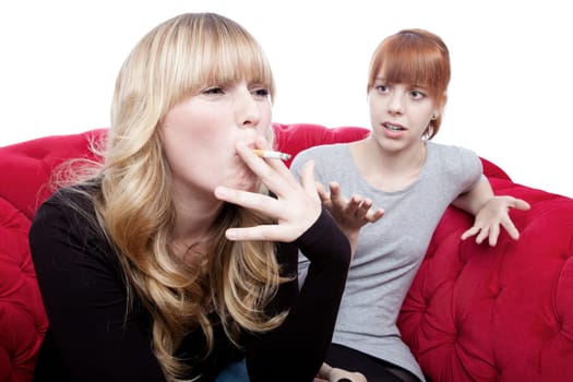 young beautiful blond and red haired girls is shocked about friend smoking on red sofa in front of white background