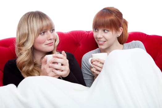 young beautiful blond and red haired girls on red sofa with coffee under blanket in front of white background