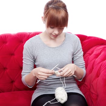 young beautiful red haired girl knitting on red sofa in front of white background