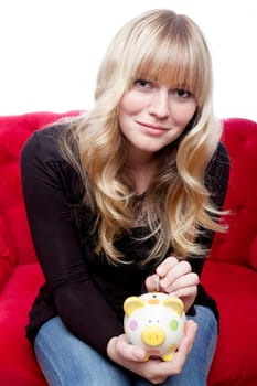young blond haired girl putting coin in piggybank on red sofa in front of white background
