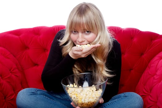 young blond haired girl eat a hand of popcorn on red sofa in front of white background
