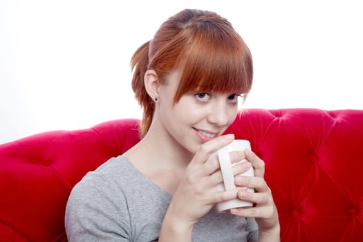 young beautiful red haired girl on red sofa with cup of tea in front of white background