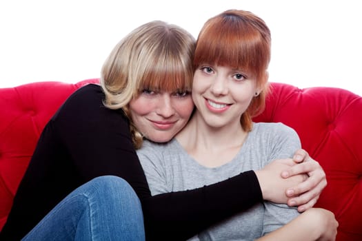young beautiful blond and red haired girls hug while sitting on red sofa in front of white background